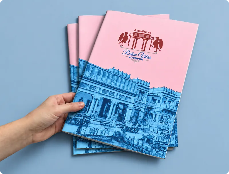 Stack of Ratan Villa menus with a pink cover and a blue sketch of the palace, held in one hand